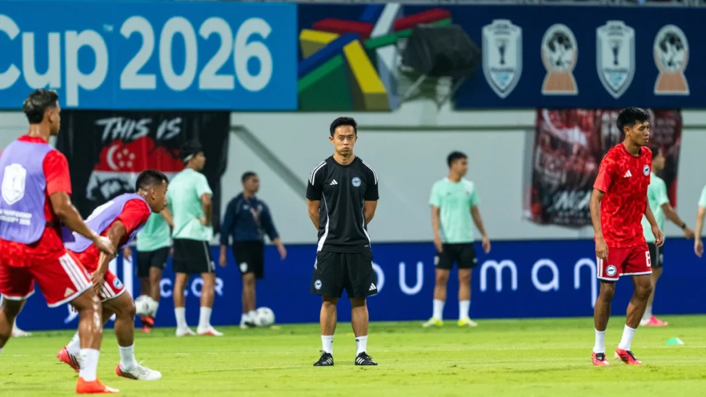 Coach in black kit stands with hands behind back on a soccer field, surrounded by players in red and mint training vests during a practice session under a'Cup 2026' banner