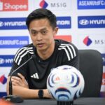 Man in a black Adidas shirt sits at a press conference table with a microphone, a white-blue-red soccer ball in front, and sponsor logos on the backdrop.