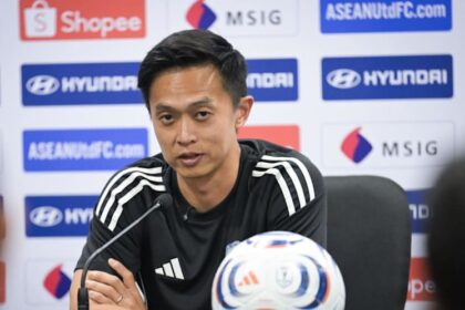 Man in a black Adidas shirt sits at a press conference table with a microphone, a white-blue-red soccer ball in front, and sponsor logos on the backdrop.
