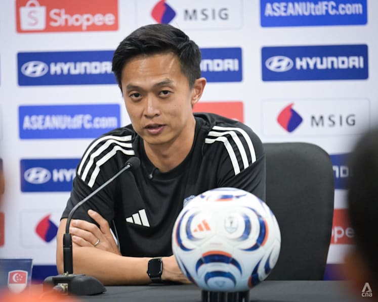 Man in a black Adidas shirt sits at a press conference table with a microphone, a white-blue-red soccer ball in front, and sponsor logos on the backdrop.