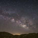 Milky Way arc across a starry night sky over rocky desert hills, with a meteor streaking through.