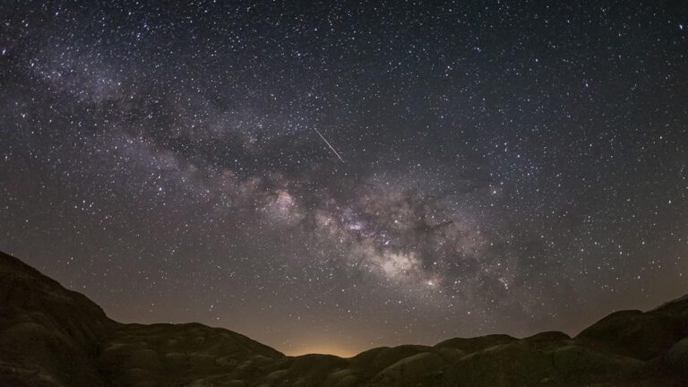 Milky Way arc across a starry night sky over rocky desert hills, with a meteor streaking through.