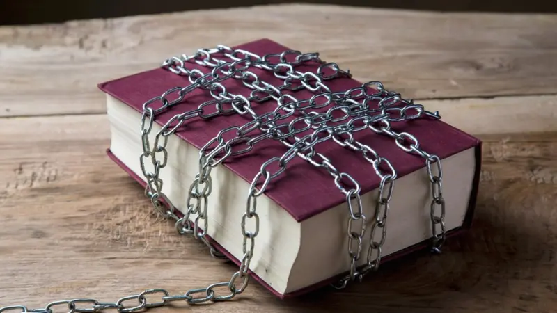 A thick hardcover book bound with heavy metal chains sprawled on a wooden table, symbolizing censorship or restriction.