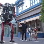 Man in a black shirt stands beside a towering red mechanical robot on a city street, with two smaller robots nearby.