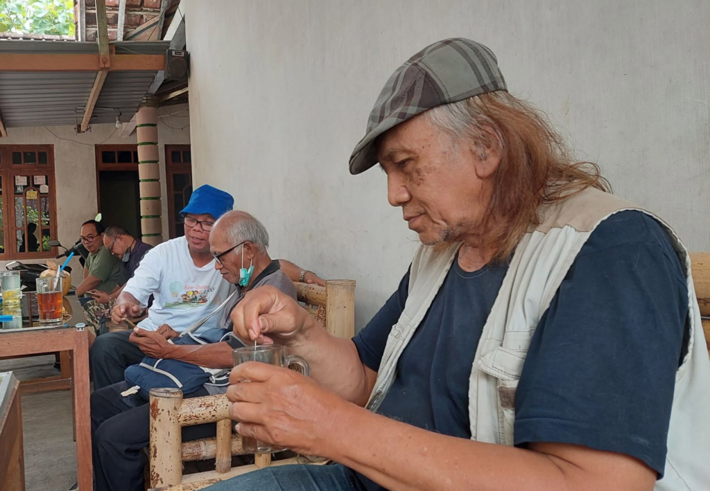 A group of elderly men sit at a wooden table on a porch, drinking and chatting; man in foreground wears a plaid cap and beige vest, stirring a glass with a straw.