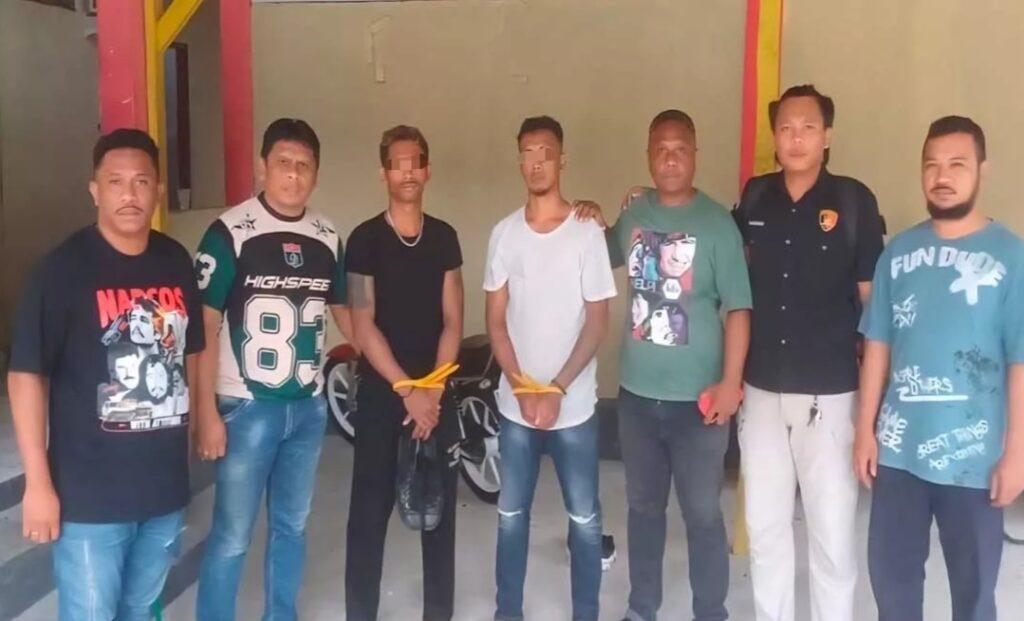 Eight men stand in a row indoors; several have yellow restraints around their wrists or hands tied with yellow bands.