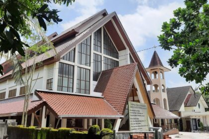 Modern campus building with red-tiled, multi-gabled roof and a tall arched bell tower, set among lush trees and a blue sky.