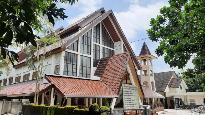 Modern campus building with red-tiled, multi-gabled roof and a tall arched bell tower, set among lush trees and a blue sky.