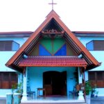 Blue-walled church with a steep red-tiled roof, open doors, and a cross atop the peak; entrance area with benches and plants.
