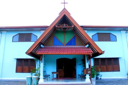 Blue-walled church with a steep red-tiled roof, open doors, and a cross atop the peak; entrance area with benches and plants.