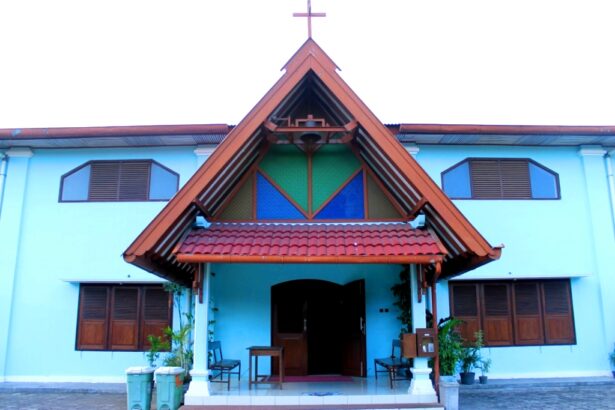 Blue-walled church with a steep red-tiled roof, open doors, and a cross atop the peak; entrance area with benches and plants.