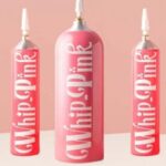Five pink cosmetic squeeze-tube bottles arranged in a row on a light pink background, labeled lip whip.