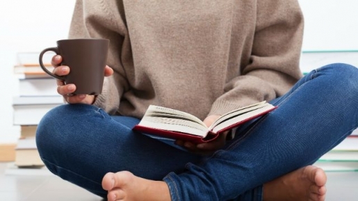 Person sitting cross-legged on the floor in a beige sweater and blue jeans, holding a mug and an open book, with stacks of books nearby for a cozy reading scene.