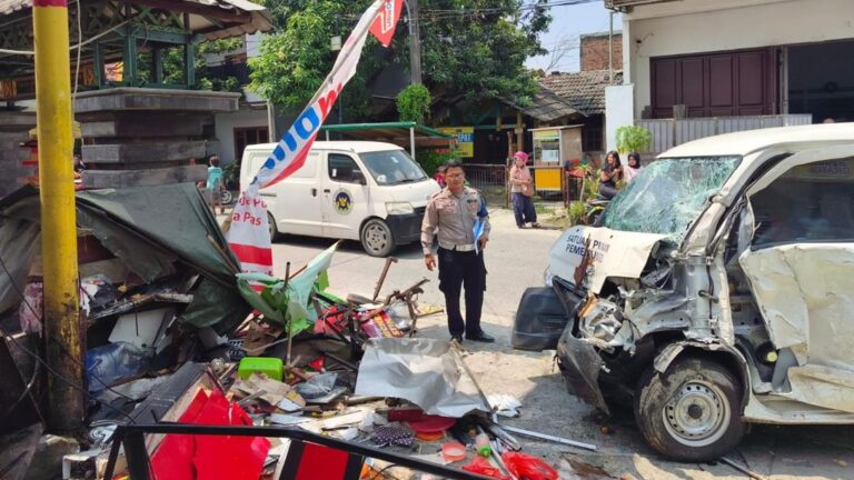Police officer in uniform stands amid a vehicle crash scene with a severely damaged white van and scattered debris on a street, bystanders in the background.