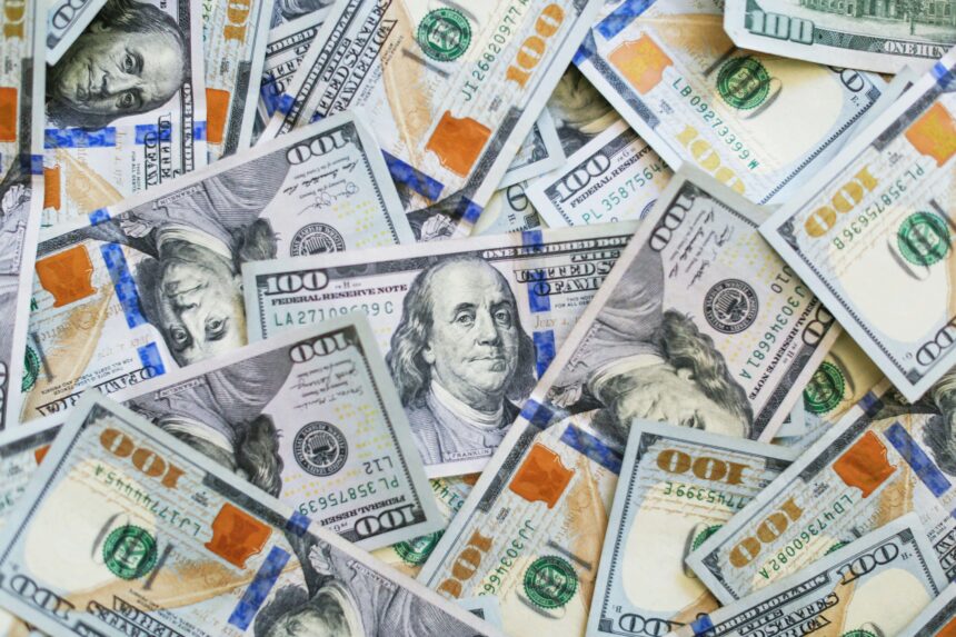 Pile of scattered US $100 bills with Benjamin Franklin portraits and blue security strips, representing cash cluttered together.