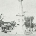 Historic street scene: a town square with a tall monument or column at a circular plaza, surrounded by small shops, trees, and early-20th-century traffic including bicycles and cars.