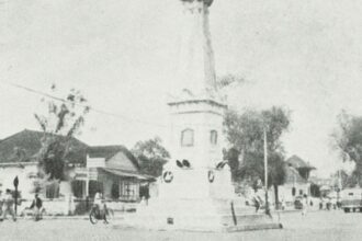 Historic street scene: a town square with a tall monument or column at a circular plaza, surrounded by small shops, trees, and early-20th-century traffic including bicycles and cars.