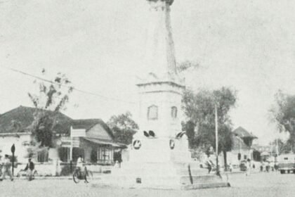 Historic street scene: a town square with a tall monument or column at a circular plaza, surrounded by small shops, trees, and early-20th-century traffic including bicycles and cars.