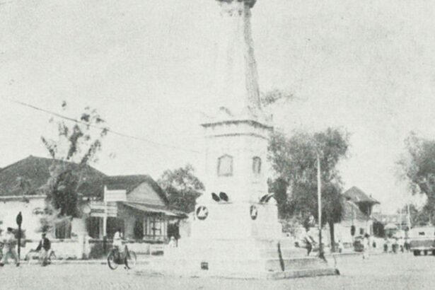 Historic street scene: a town square with a tall monument or column at a circular plaza, surrounded by small shops, trees, and early-20th-century traffic including bicycles and cars.