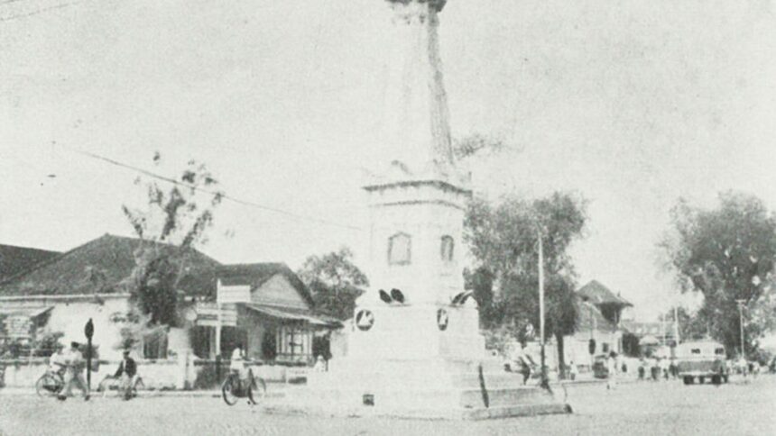 Historic street scene: a town square with a tall monument or column at a circular plaza, surrounded by small shops, trees, and early-20th-century traffic including bicycles and cars.