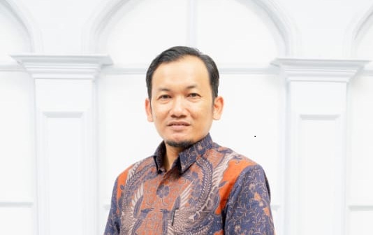 Portrait of a man in a patterned batik shirt posing indoors against a bright white architectural backdrop.