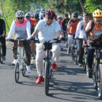 Group of cyclists riding together on a rural road, front row wearing helmets and colorful jerseys fallowing a lead rider in white polo.