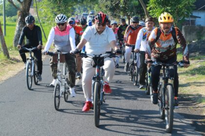 Group of cyclists riding together on a rural road, front row wearing helmets and colorful jerseys fallowing a lead rider in white polo.