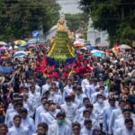 A large crowd in a street parade, with white-clad participants carrying a tall, green-yellow ceremonial structure atop a blue cloth, surrounded by spectators and umbrellas.