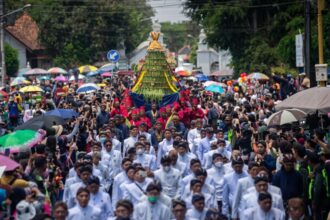 A large crowd in a street parade, with white-clad participants carrying a tall, green-yellow ceremonial structure atop a blue cloth, surrounded by spectators and umbrellas.