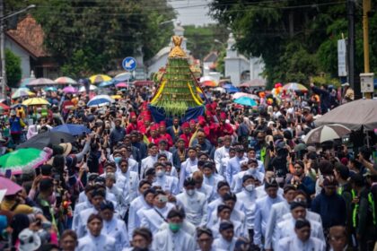 A large crowd in a street parade, with white-clad participants carrying a tall, green-yellow ceremonial structure atop a blue cloth, surrounded by spectators and umbrellas.