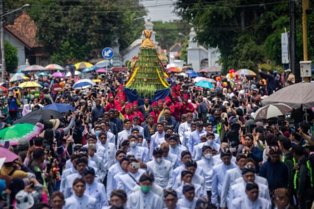 A large crowd in a street parade, with white-clad participants carrying a tall, green-yellow ceremonial structure atop a blue cloth, surrounded by spectators and umbrellas.