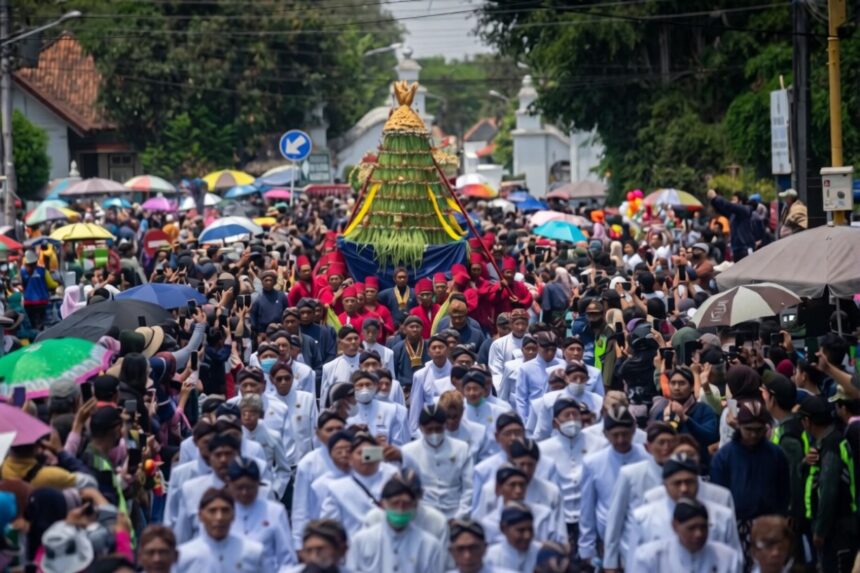 A large crowd in a street parade, with white-clad participants carrying a tall, green-yellow ceremonial structure atop a blue cloth, surrounded by spectators and umbrellas.