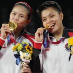 Two female athletes in matching red‑white jackets celebrate on a podium, biting their gold medals and holding bouquets with sunflowers.