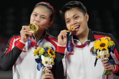 Two female athletes in matching red‑white jackets celebrate on a podium, biting their gold medals and holding bouquets with sunflowers.