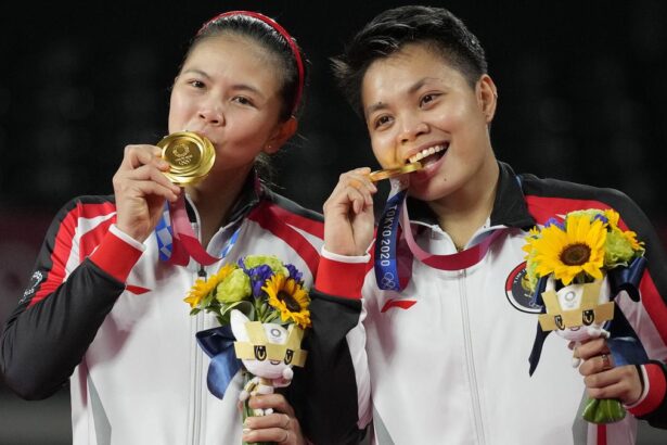 Two female athletes in matching red‑white jackets celebrate on a podium, biting their gold medals and holding bouquets with sunflowers.