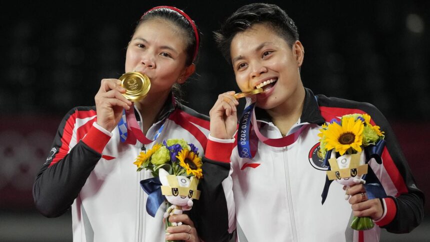 Two female athletes in matching red‑white jackets celebrate on a podium, biting their gold medals and holding bouquets with sunflowers.