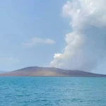 Calm blue sea with a flat volcanic island in the distance and white smoke rising from a crater.