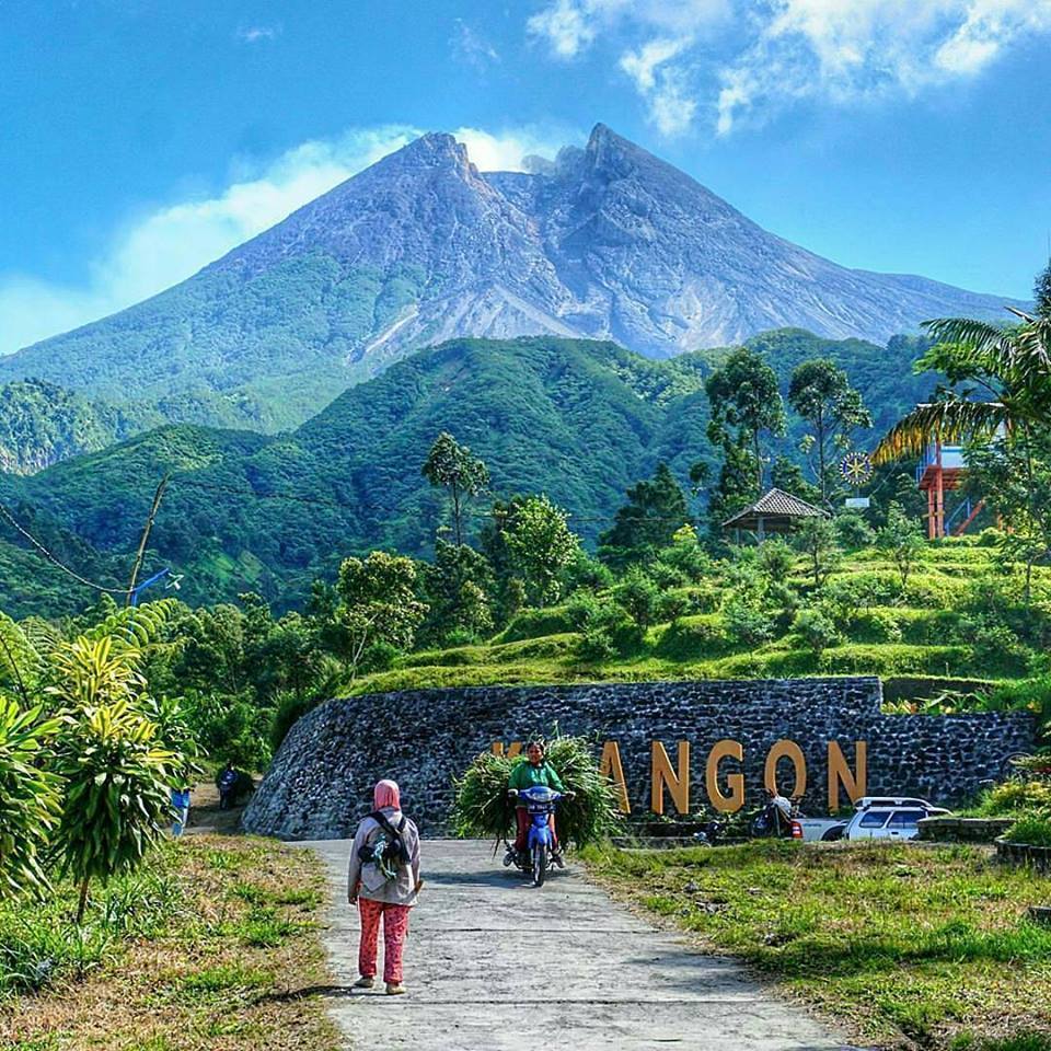 Volcano mountain rising above green hills, with a stone wall sign reading 'ANGON' and people walking along a path.