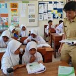 In a classroom, a male teacher in a beige uniform hands out papers to a group of girls wearing white hijabs as they read textbooks at wooden desks.