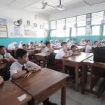 A classroom full of students in white shirts seated at wooden desks, facing a teacher at the front of the room.