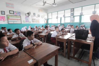 A classroom full of students in white shirts seated at wooden desks, facing a teacher at the front of the room.