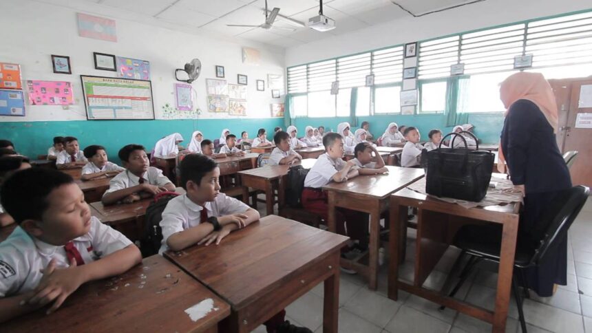 guru mengajar di kelas - Mabur.co A classroom full of students in white shirts seated at wooden desks, facing a teacher at the front of the room.