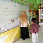 Teacher in a beige hijab explains math problems on a whiteboard to a young student in a colorful classroom.
