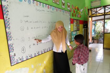 Teacher in a beige hijab explains math problems on a whiteboard to a young student in a colorful classroom.