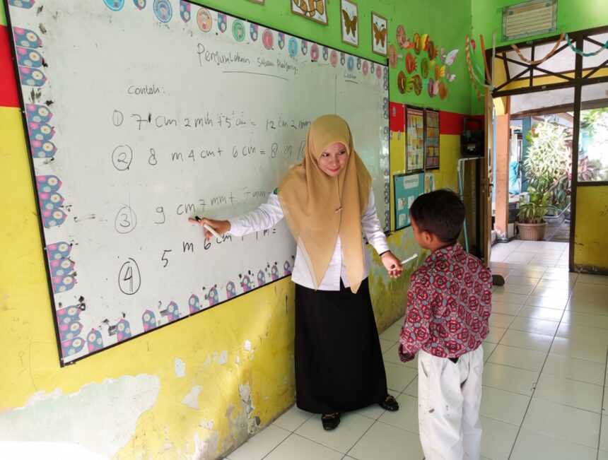 Teacher in a beige hijab explains math problems on a whiteboard to a young student in a colorful classroom.