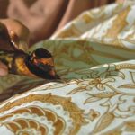 Close-up of a hand applying hot wax to batik fabric, with a brown patterned cloth in the background.