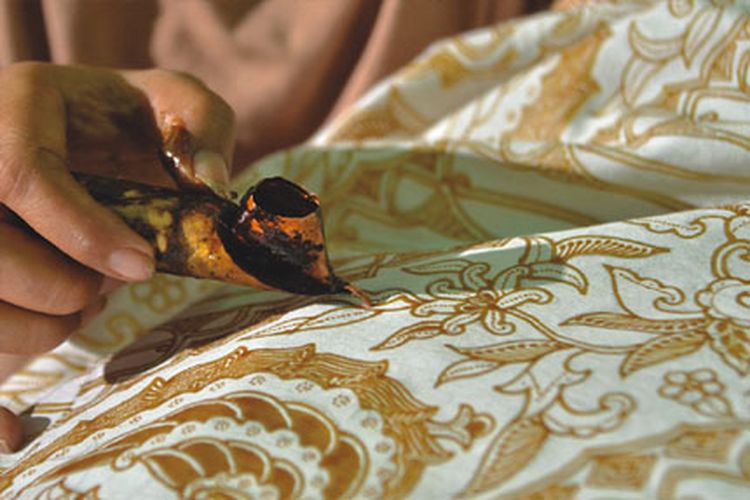 Close-up of a hand applying hot wax to batik fabric, with a brown patterned cloth in the background.