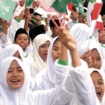 Group of smiling girls in white hijabs waving green and white flags at a rally.
