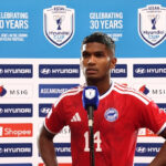 Soccer player in a red jersey with number 14 speaks at a press conference, blue backdrop showing ASEAN Cup logos and sponsors behind him.