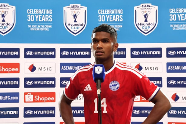 Soccer player in a red jersey with number 14 speaks at a press conference, blue backdrop showing ASEAN Cup logos and sponsors behind him.